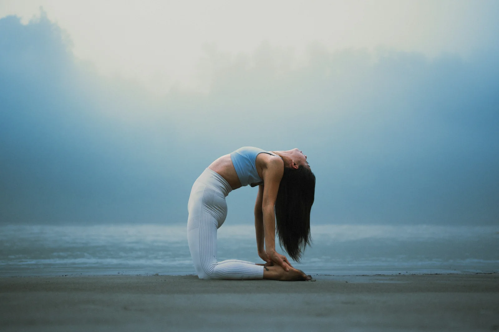 A person holds a yoga balance pose on a beach beside the water.