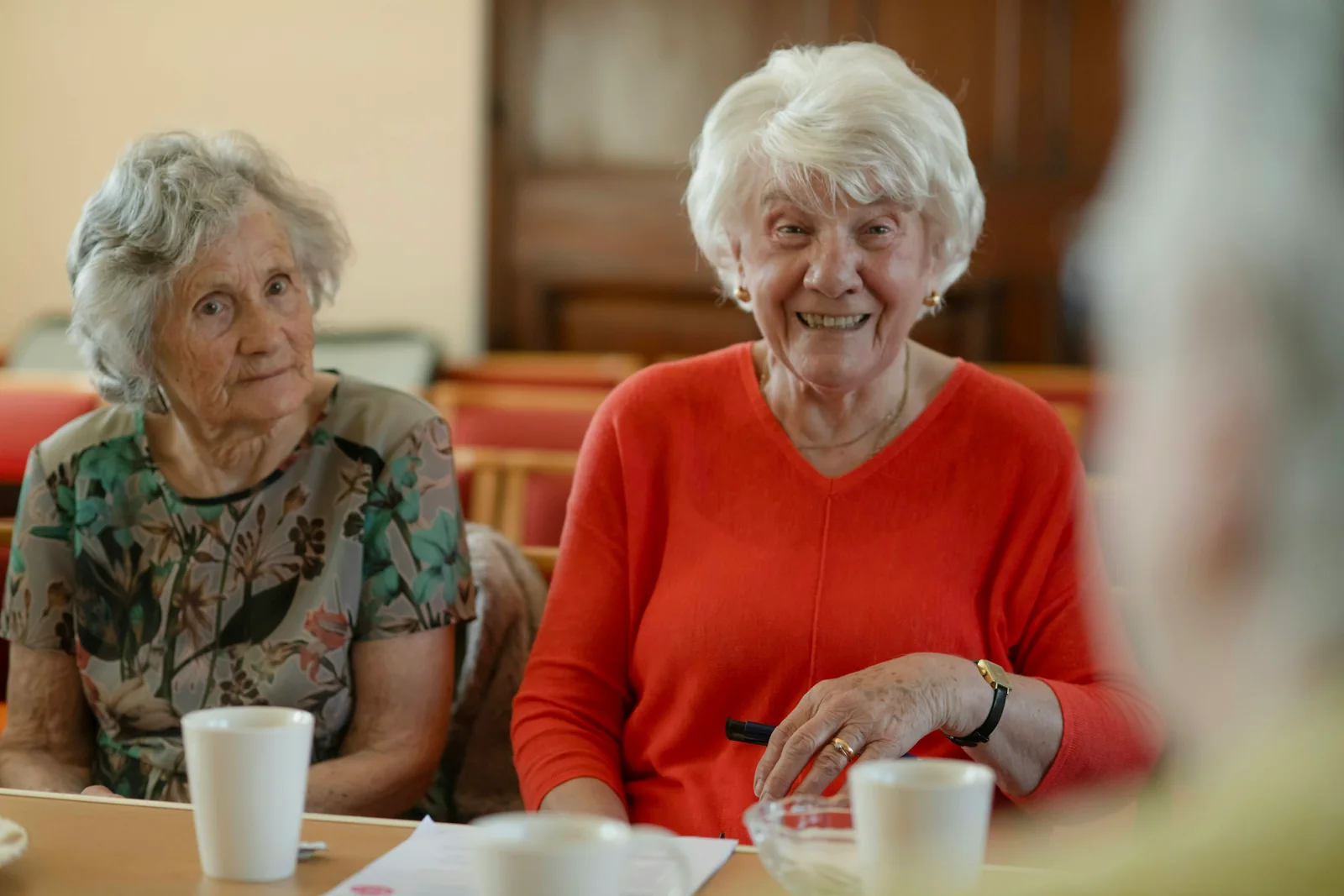 Two older women sit at a table, one smiling toward the camera.